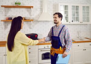 A residential plumber, wearing a uniform with the company logo, is shaking hands with a satisfied homeowner.